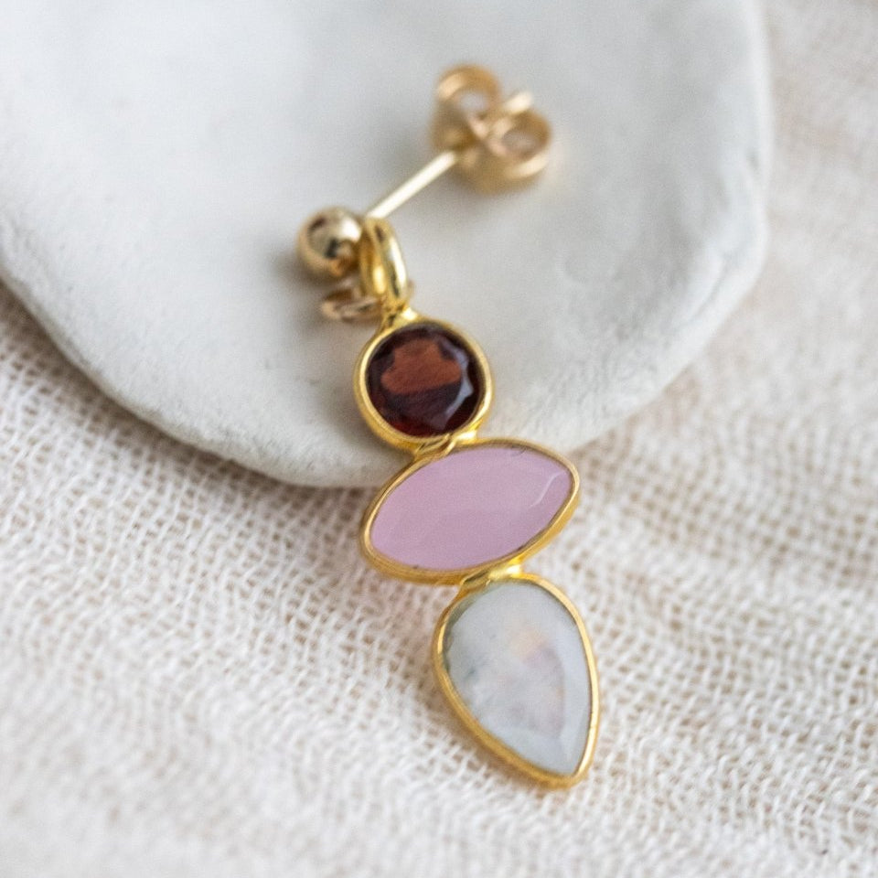 Ein goldener Multi-Edelstein-Ohrstecker, bestehend aus Granat, rosa Chalcedon und Mondstein, lehnt an einem weißen Schälchen.