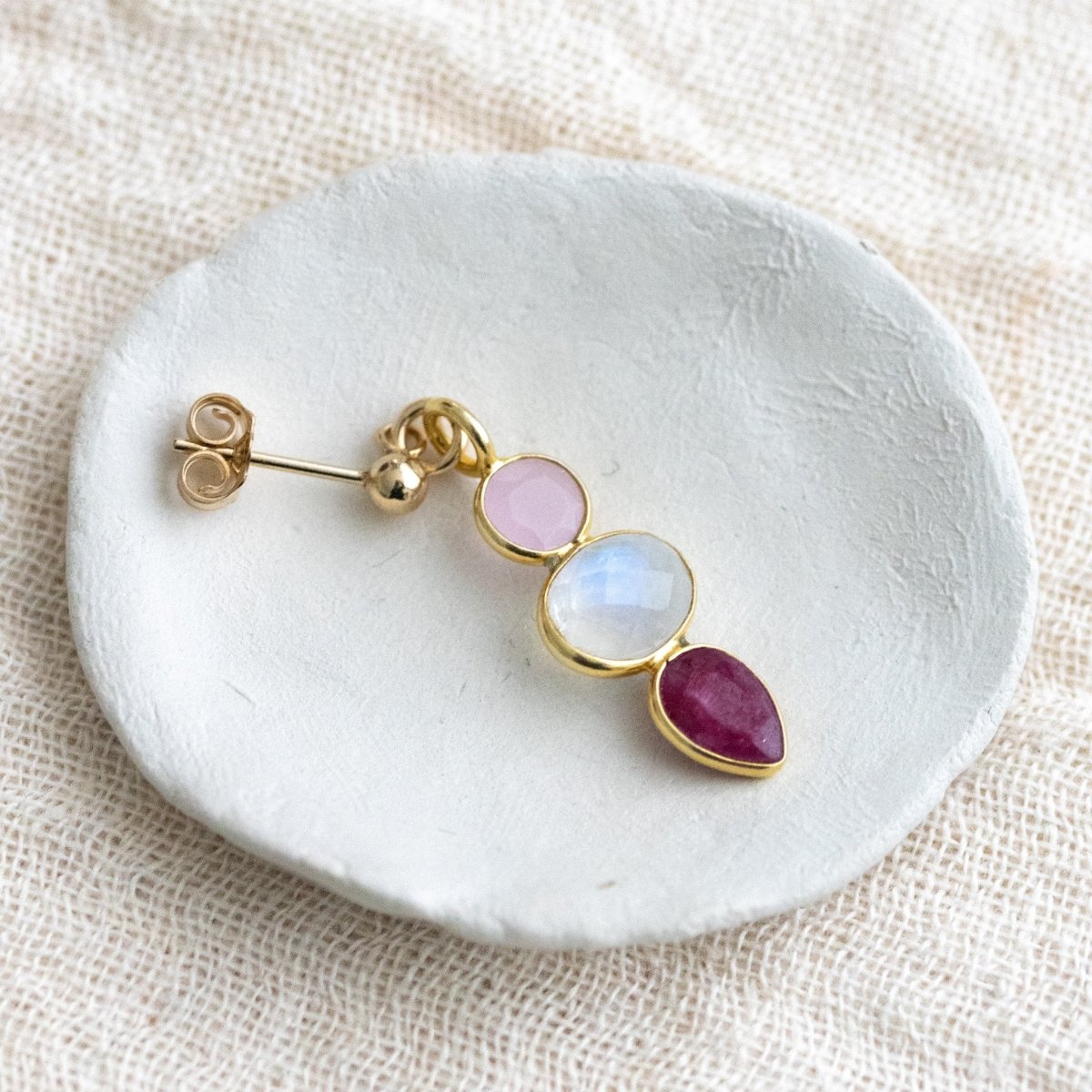 Ein goldener Ohrstecker mit einem rosa Chalcedon, einem Mondstein und einem Rubin liegt auf einem weißen Schälchen.