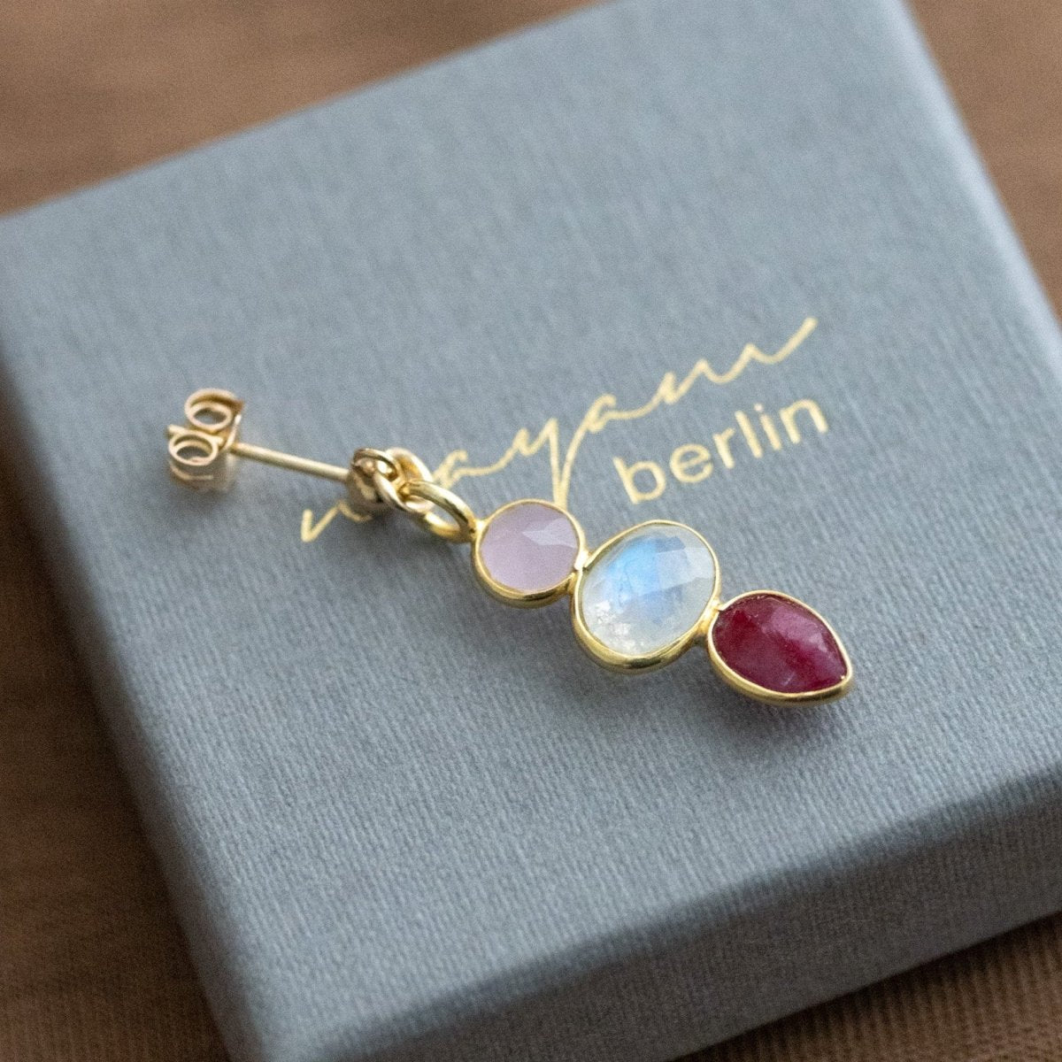 Ein goldener Ohrstecker mit einem rosa Chalcedon, einem Mondstein und einem Rubin liegt auf einem grauen Schmucketui.