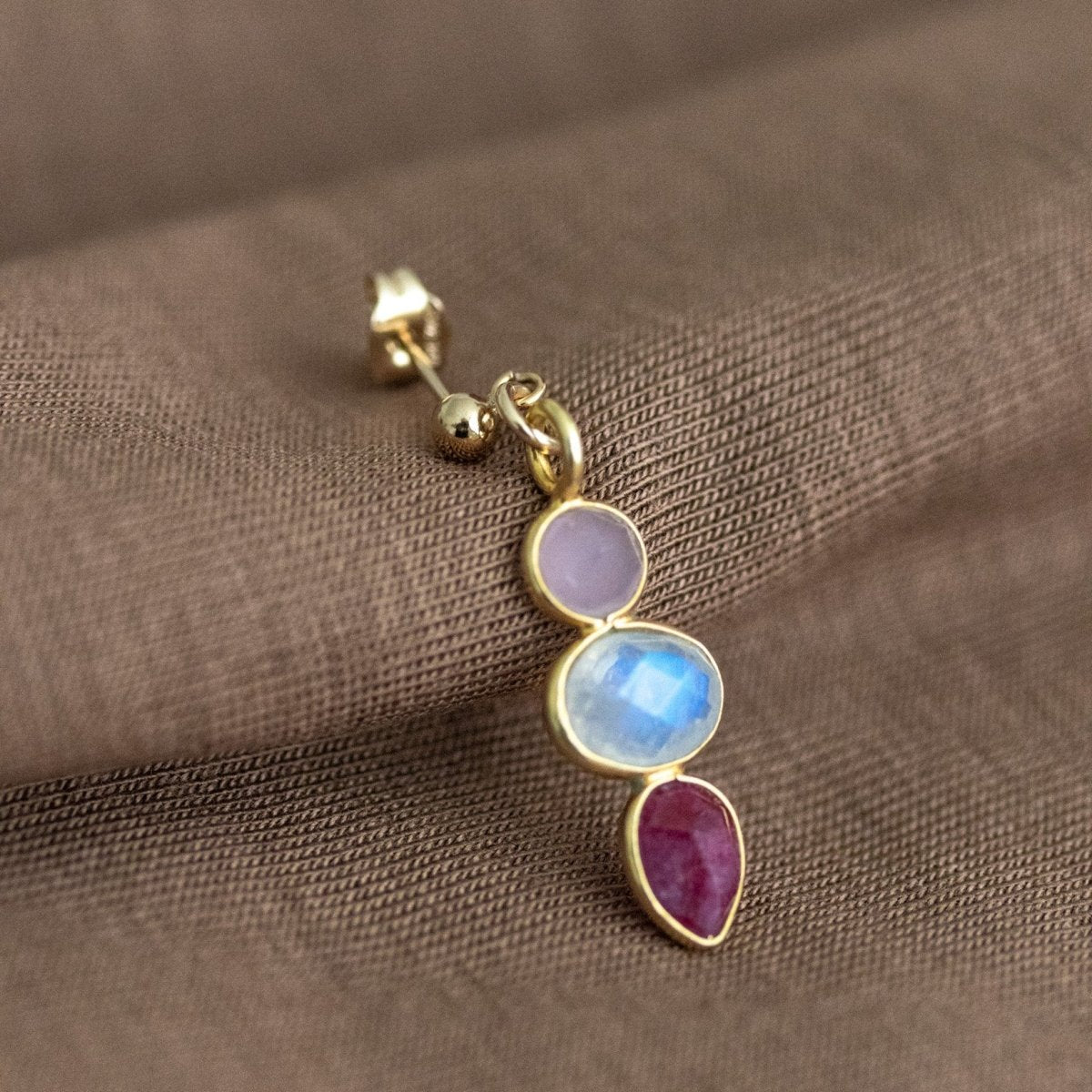 Ein goldener Ohrstecker mit einem rosa Chalcedon, einem Mondstein und einem Rubin lehnt an braunem Stoff.