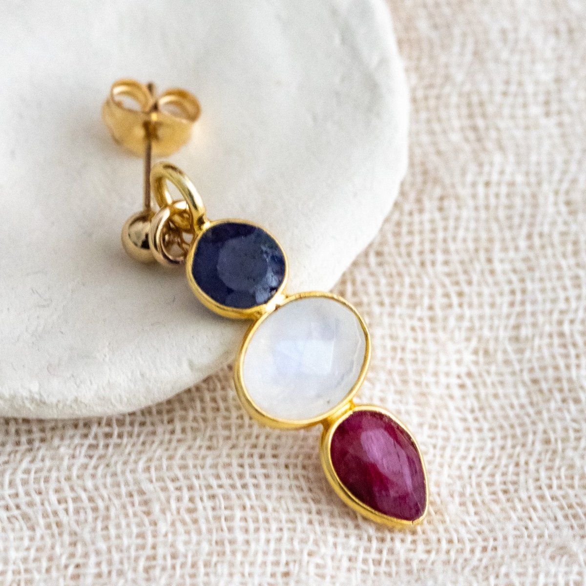 Ein Multi-Edelstein Ohrring mit Saphir, Mondstein und Rubin in gold gefasst lehnt an einem weißen Schälchen auf cremefarbenen Leinenstoff.