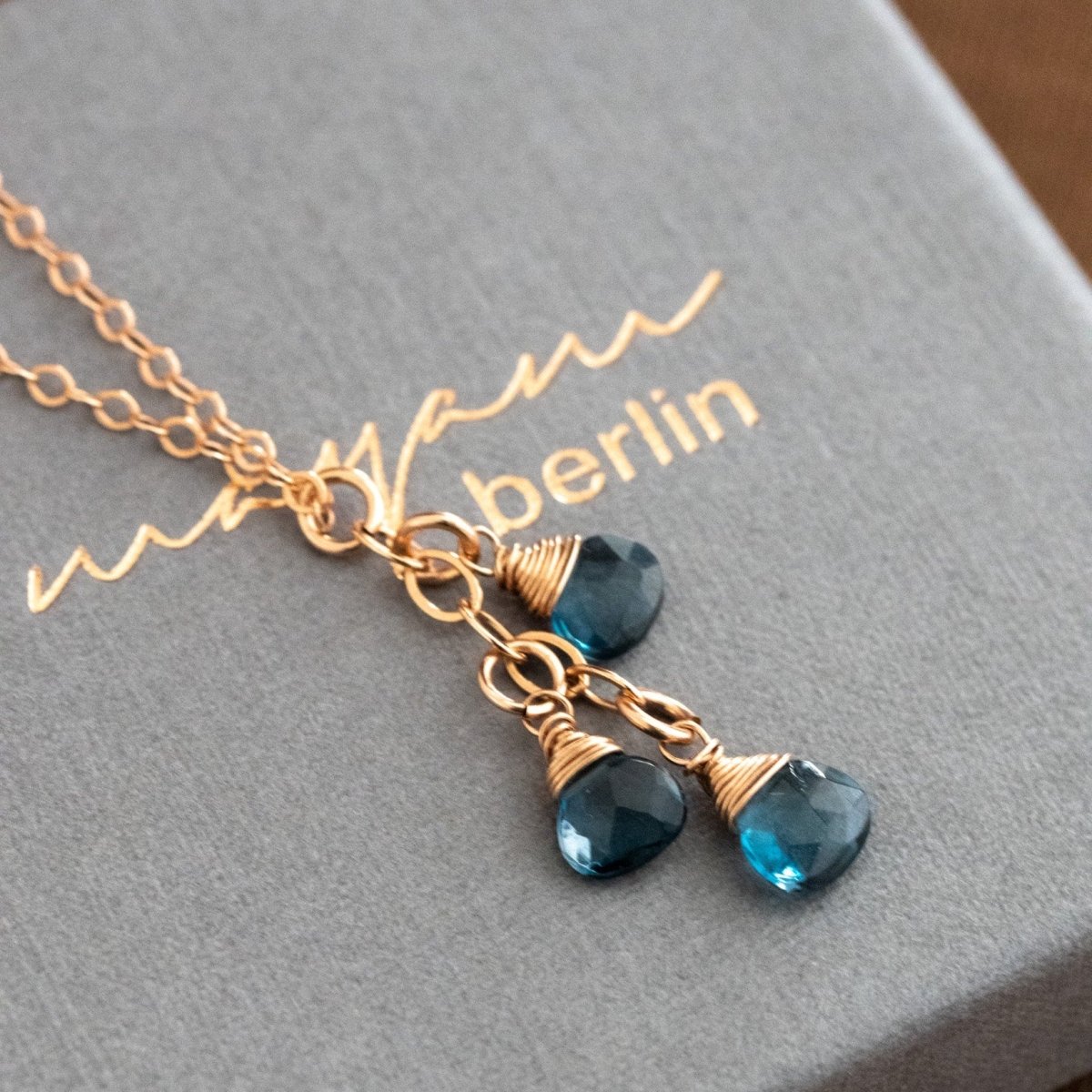 Auf einem grauen Schmuckkarton liegt eine feine Roségold-Kette mit drei kleinen, untereinander hängenden London Blautopas-Tropfen.