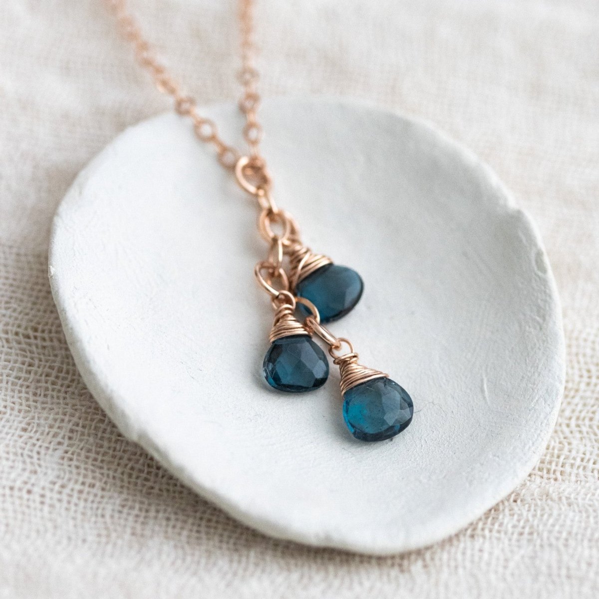 Auf einem weißen Schälchen liegt eine feine Roségold-Kette mit drei kleinen, untereinander hängenden London Blautopas-Tropfen.