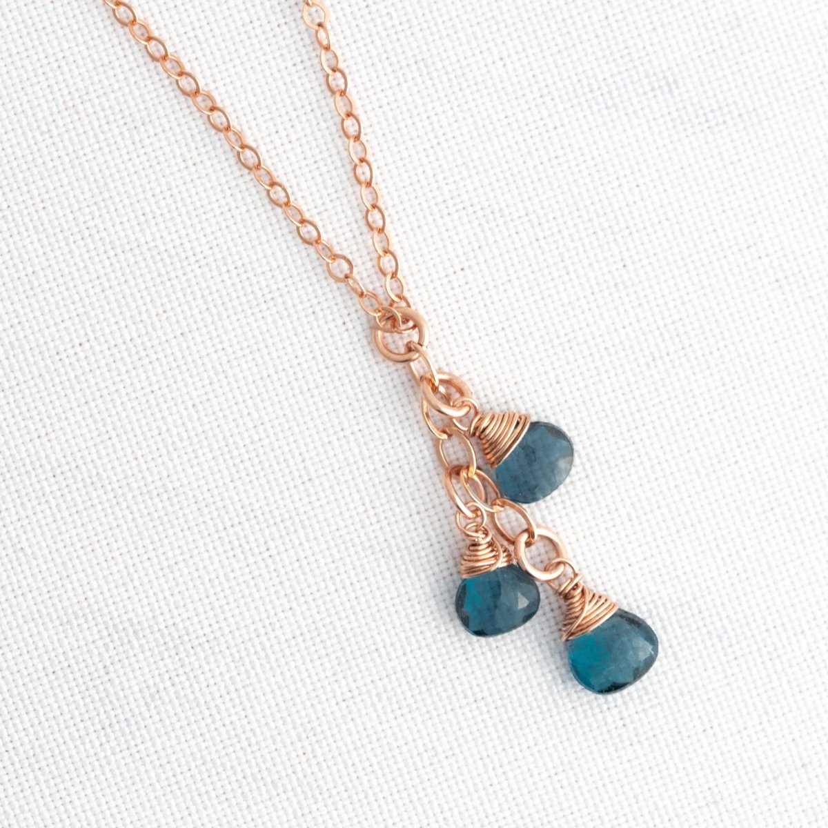 Vor weißem Hintergrund hängt eine feine Roségold-Kette mit drei kleinen, untereinander hängenden London Blautopas-Tropfen.
