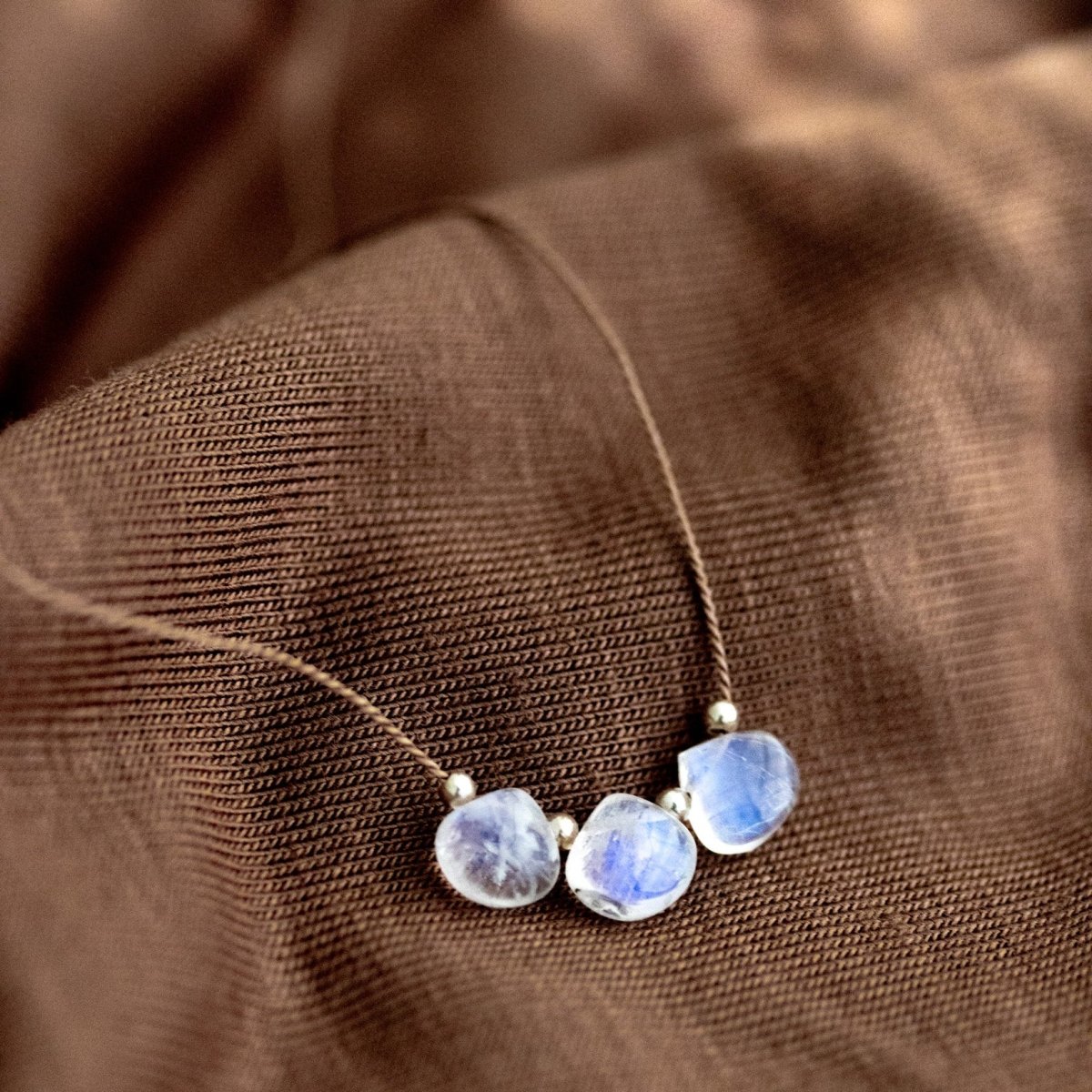 Eine dünne Seidenkette mit drei aufgezogenen Mondsteinen liegt auf braunem Stoffuntergrund.
