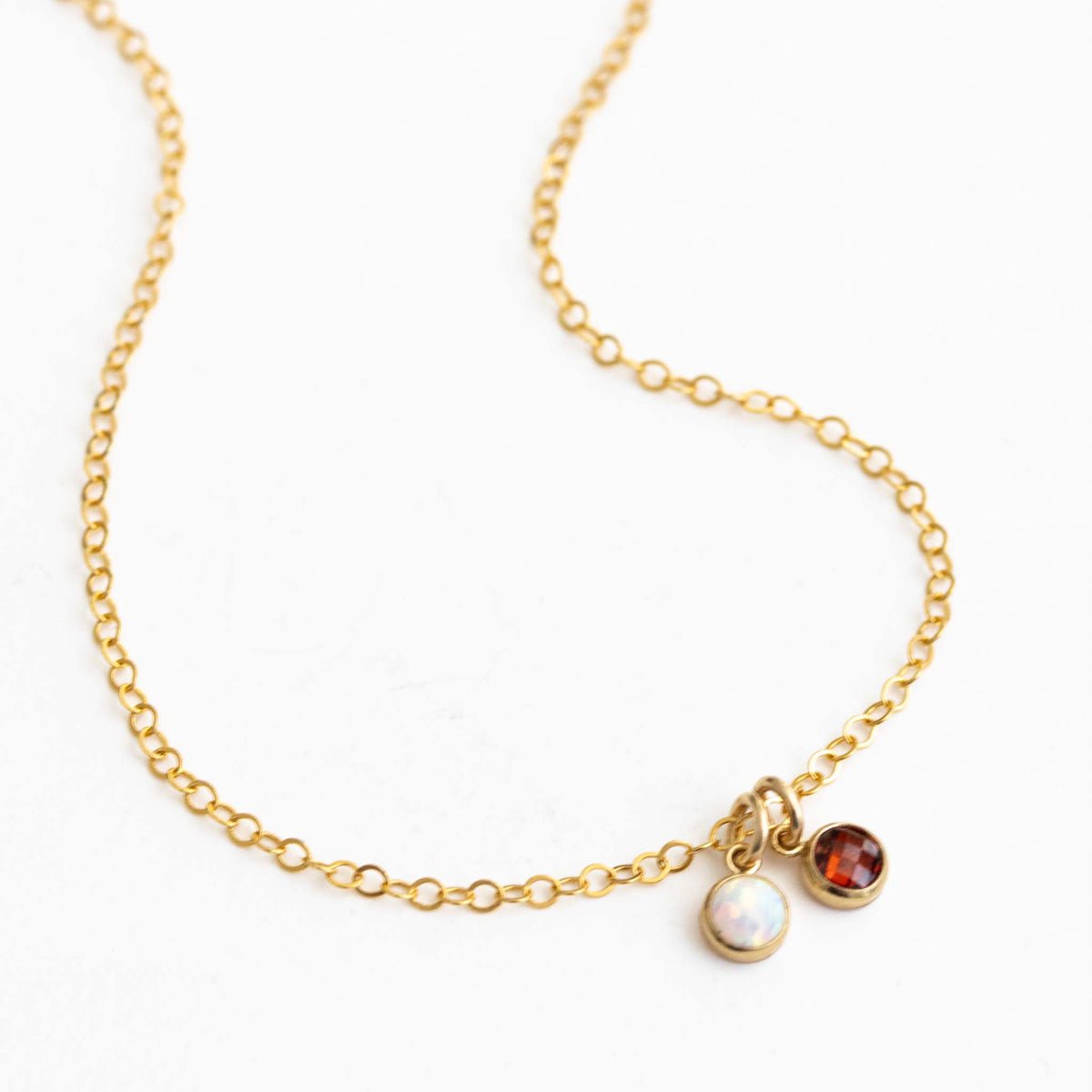 Eine zierliche Goldkette mit zwei winzigen, runden Anhängern, einem Opal und einem Granat, auf weißem Hintergrund.
