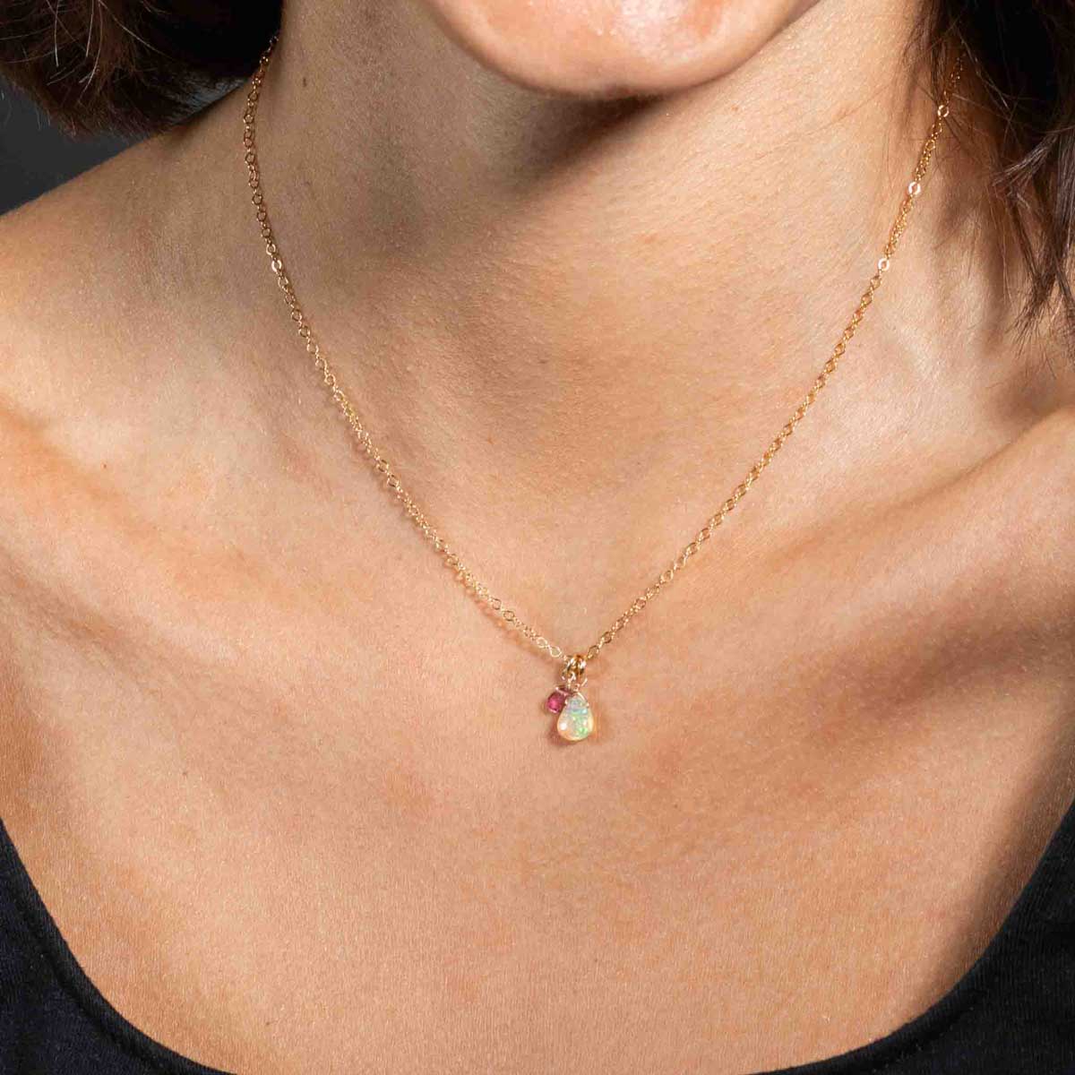 Eine zierliche Goldkette mit einem tropfenförmigen Welo Opal-Anhänger und einem winzigen rosa Turmalin hängt am Hals des Models.