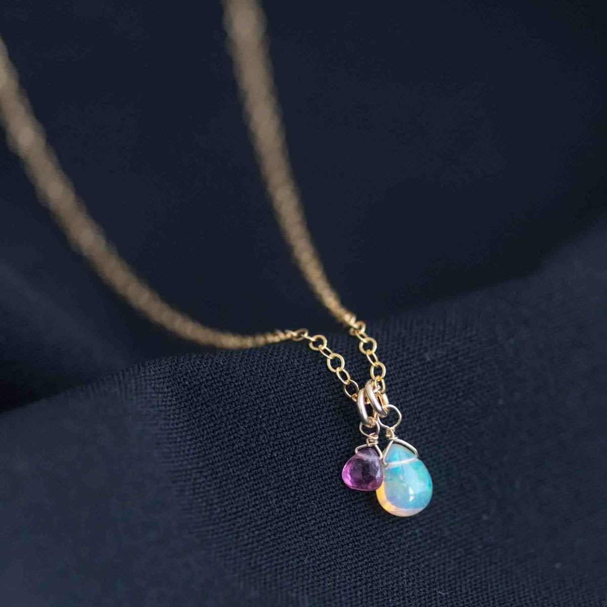 Eine zierliche Goldkette mit einem tropfenförmigen Welo Opal-Anhänger und einem winzigen rosa Turmalin liegt auf schwarzem Hintergrund.