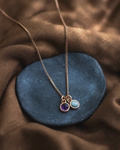 Eine feine rosé goldene Halskette mit einem Amethyst und einem Aquamarin-Anhänger liegt auf einem grauen Steinschälchen auf braunem Untergrund.