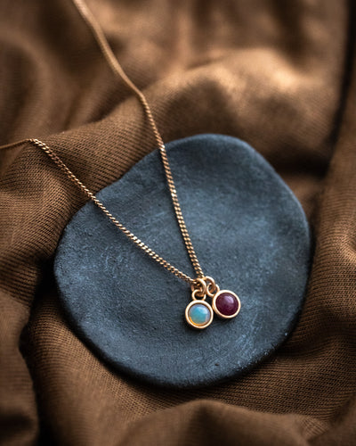 Eine feine Halskette aus rosé vergoldetem Silber mit zwei Edelstein-Anhängern, einem Opal und einem Rubin, auf einer grauen Steinschale.