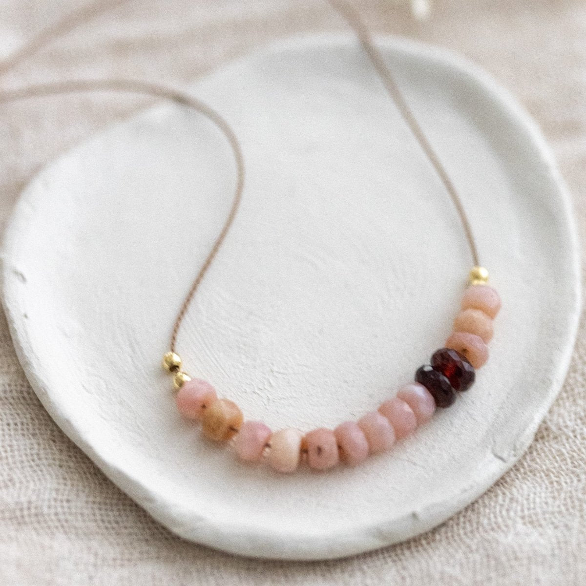 Eine Halskette aus dünner, beiger Seidenschnur, auf die eine Reihe rosa Opale und dunkelroter Granate aufgezogen sind, liegt auf einem weißen Schälchen.