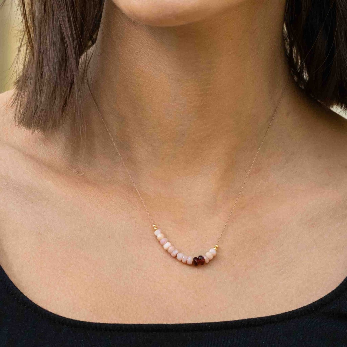 Am Hals des Models hängt eine kurze Halskette, die aus einer dünnen, beigen Seidenschnur besteht, auf die eine kurze Reihe rosa Opale und zwei schwarzrote Granate aufgezogen sind.