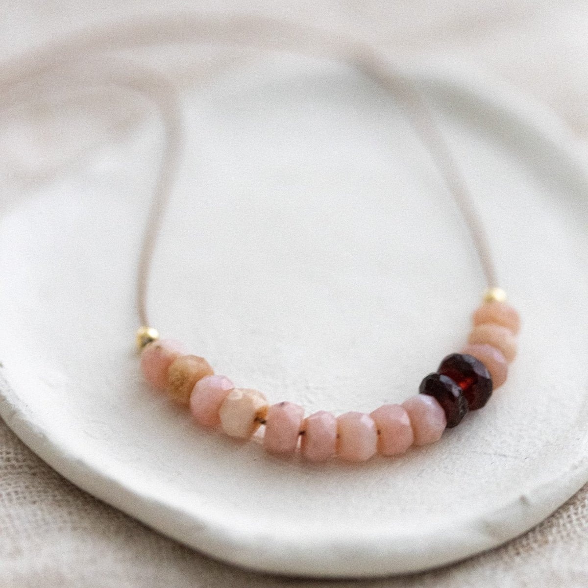 Eine Halskette aus dünner, beiger Seidenschnur, auf die eine Reihe rosa Opale und dunkelroter Granate aufgezogen sind, liegt auf einem weißen Schälchen.