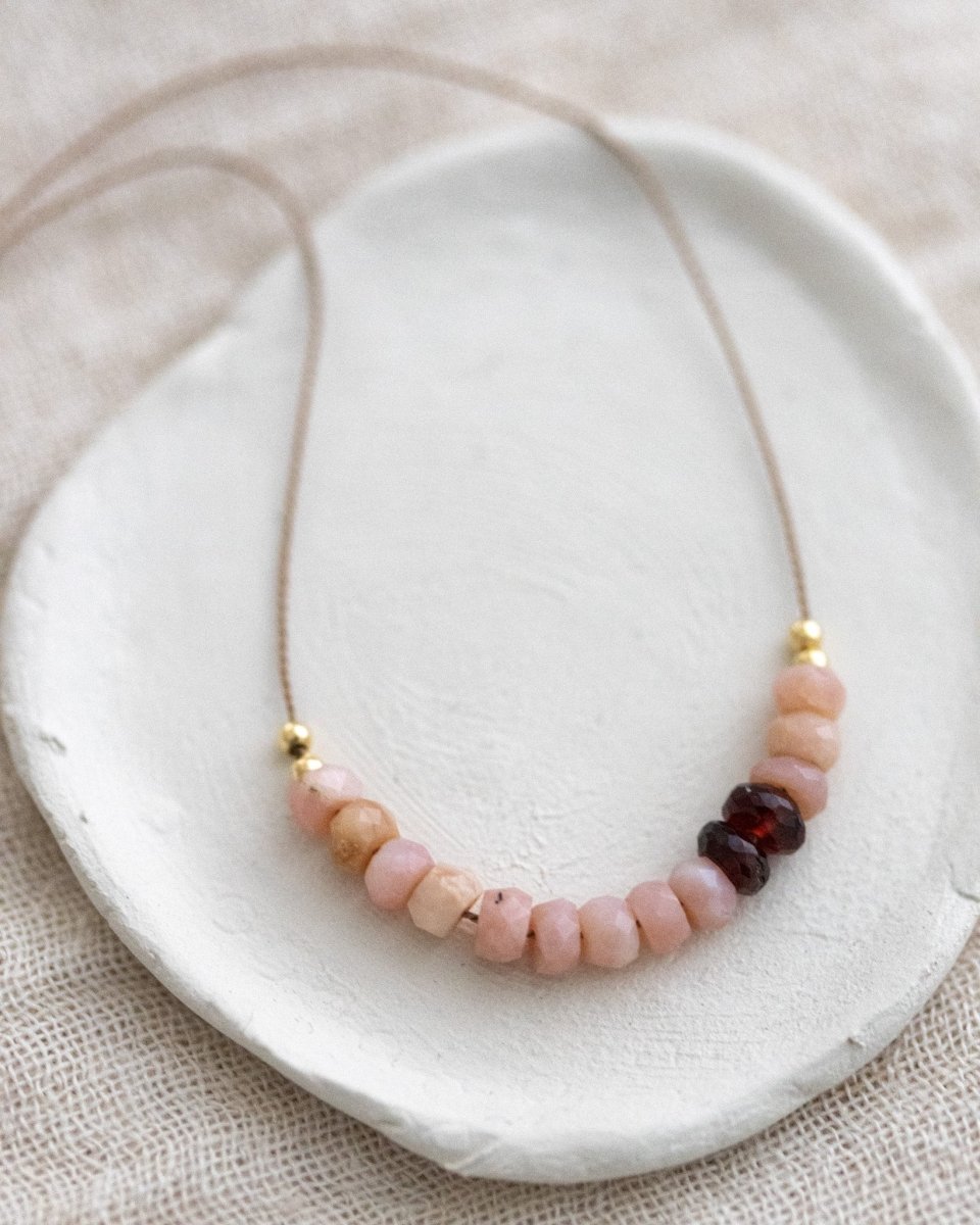 Eine Halskette aus dünner, beiger Seidenschnur, auf die eine Reihe rosa Opale und dunkelroter Granate aufgezogen sind, liegt auf einem weißen Schälchen.