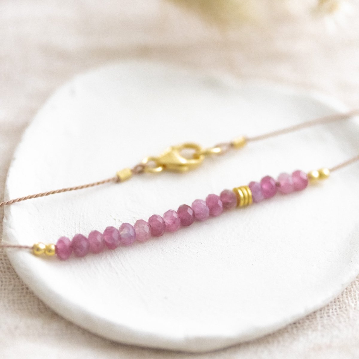 Auf weißen Schälchen liegt eine kurze Halskette. Sie besteht aus einer dünnen Seidenschnur mit einer Reihe kleiner, rosa Turmalin-Rondelle und goldenen Akzenten in der Mitte.