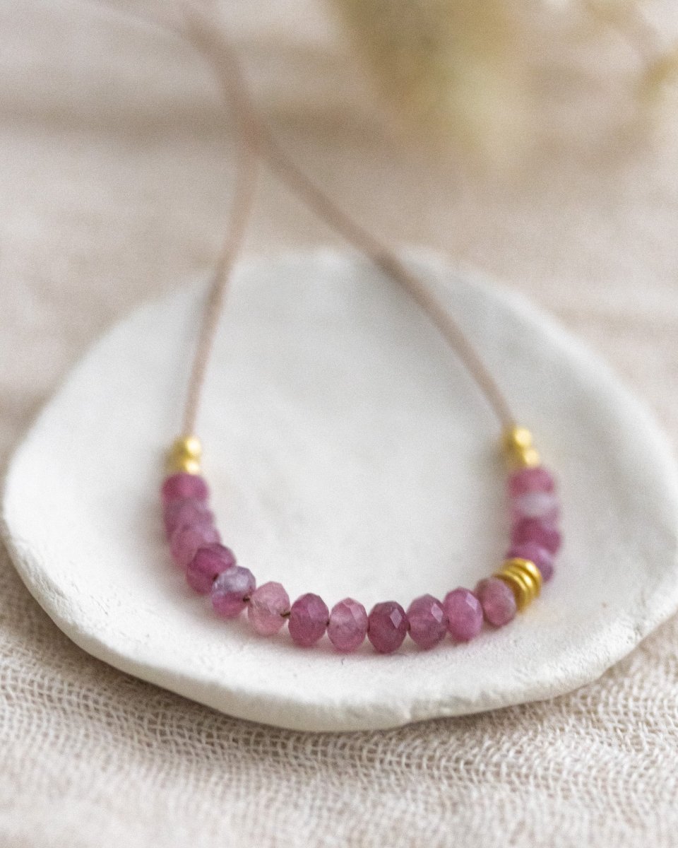 Auf weißen Schälchen liegt eine kurze Halskette. Sie besteht aus einer dünnen Seidenschnur mit einer Reihe kleiner, rosa Turmalin-Rondelle und goldenen Akzenten in der Mitte.