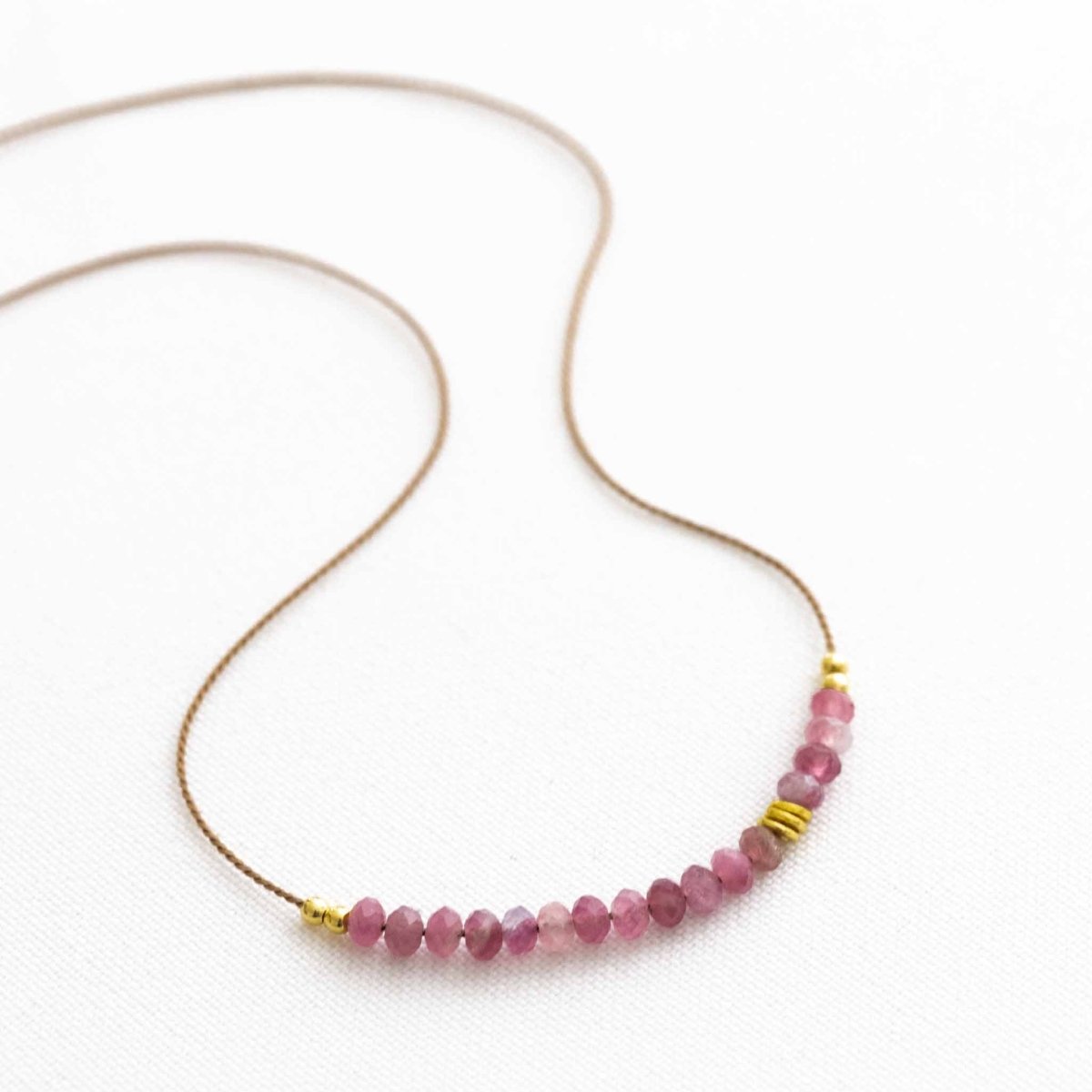 Auf weißen Hintergrund liegt eine kurze Halskette. Sie besteht aus einer dünnen Seidenschnur mit einer Reihe kleiner, rosa Turmalin-Rondelle und goldenen Akzenten in der Mitte.