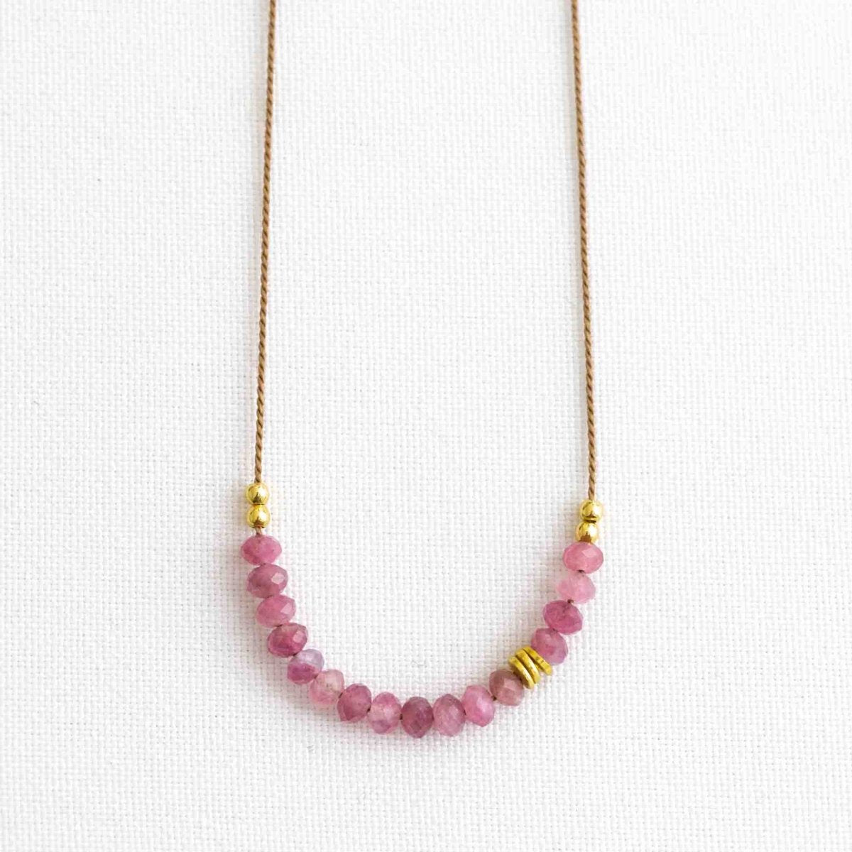 Auf weißen Hintergrund liegt eine kurze Halskette. Sie besteht aus einer dünnen Seidenschnur mit einer Reihe kleiner, rosa Turmalin-Rondelle und goldenen Akzenten in der Mitte.
