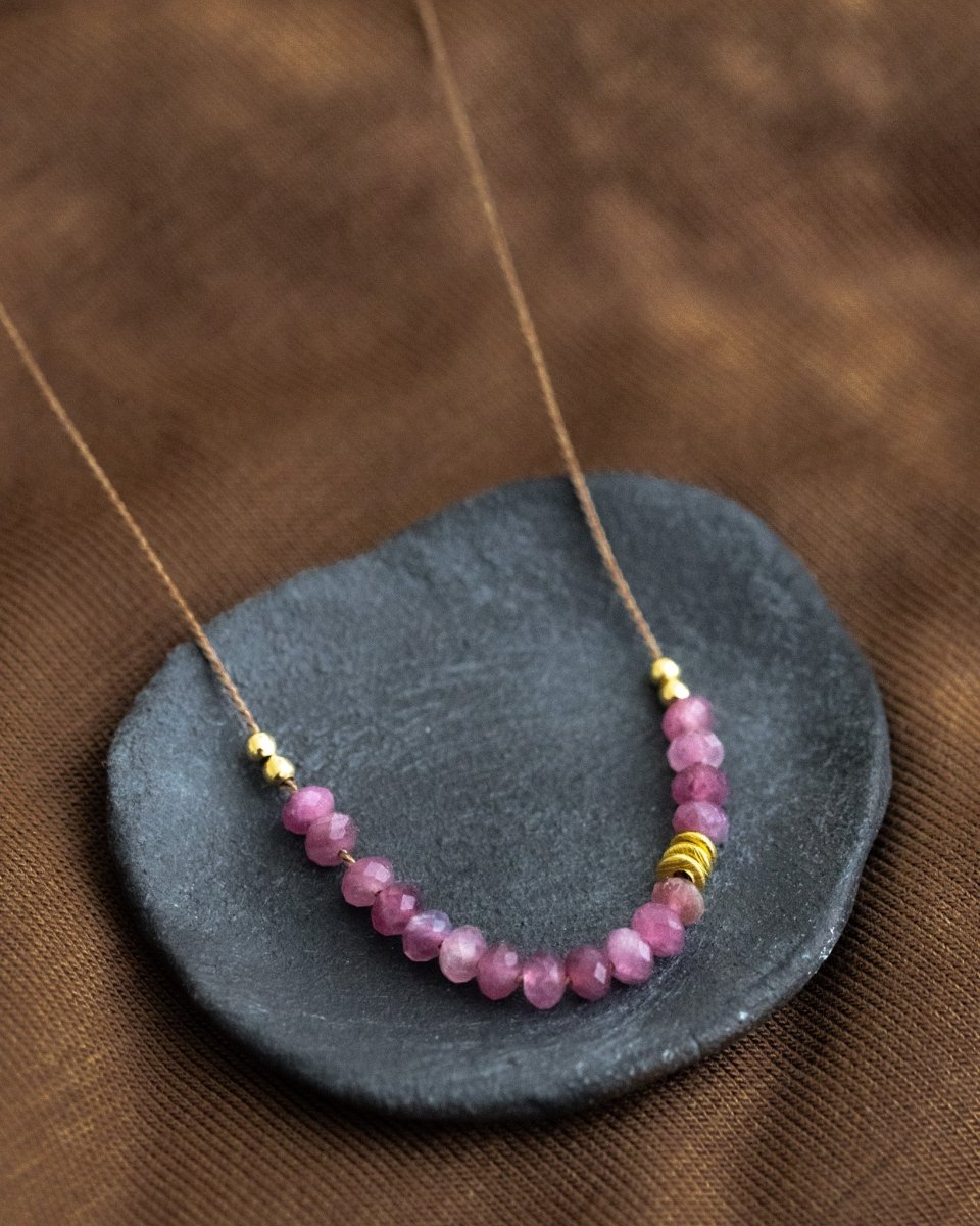 Auf dunkelgrauen Schälchen liegt eine kurze Halskette. Sie besteht aus einer dünnen Seidenschnur mit einer Reihe kleiner, rosa Turmalin-Rondelle und goldenen Akzenten in der Mitte.