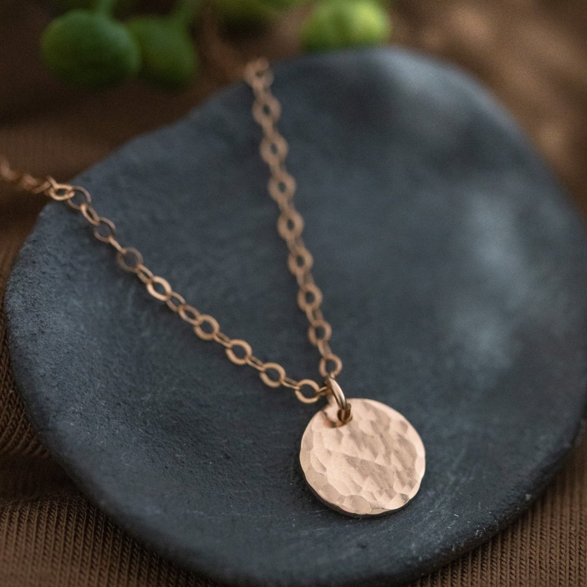 Eine rosé goldene Ankerkette mit einem runden, gehämmerten Plättchenanhänger liegt auf einem grauen Schälchen. Brauner Untergrund und ein paar verschwommene, grüne Knospen im Hintergrund.