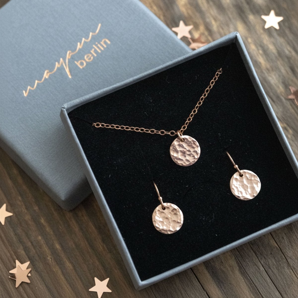 In einem offenen Schmucketui hängen eine rose goldene Kette und ein Paar rose goldener Ohrringe mit jeweils einem Plättchen. Holzuntergund mit weihnachtlichem Sternen-Konfetti.