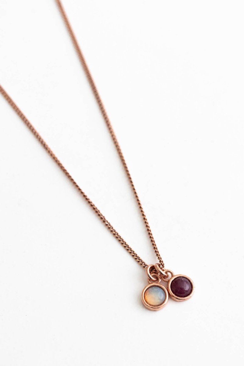 Eine feine Halskette aus rosé vergoldetem Silber mit zwei Edelstein-Anhängern, einem Opal und einem Rubin, auf weißem Hintergrund.