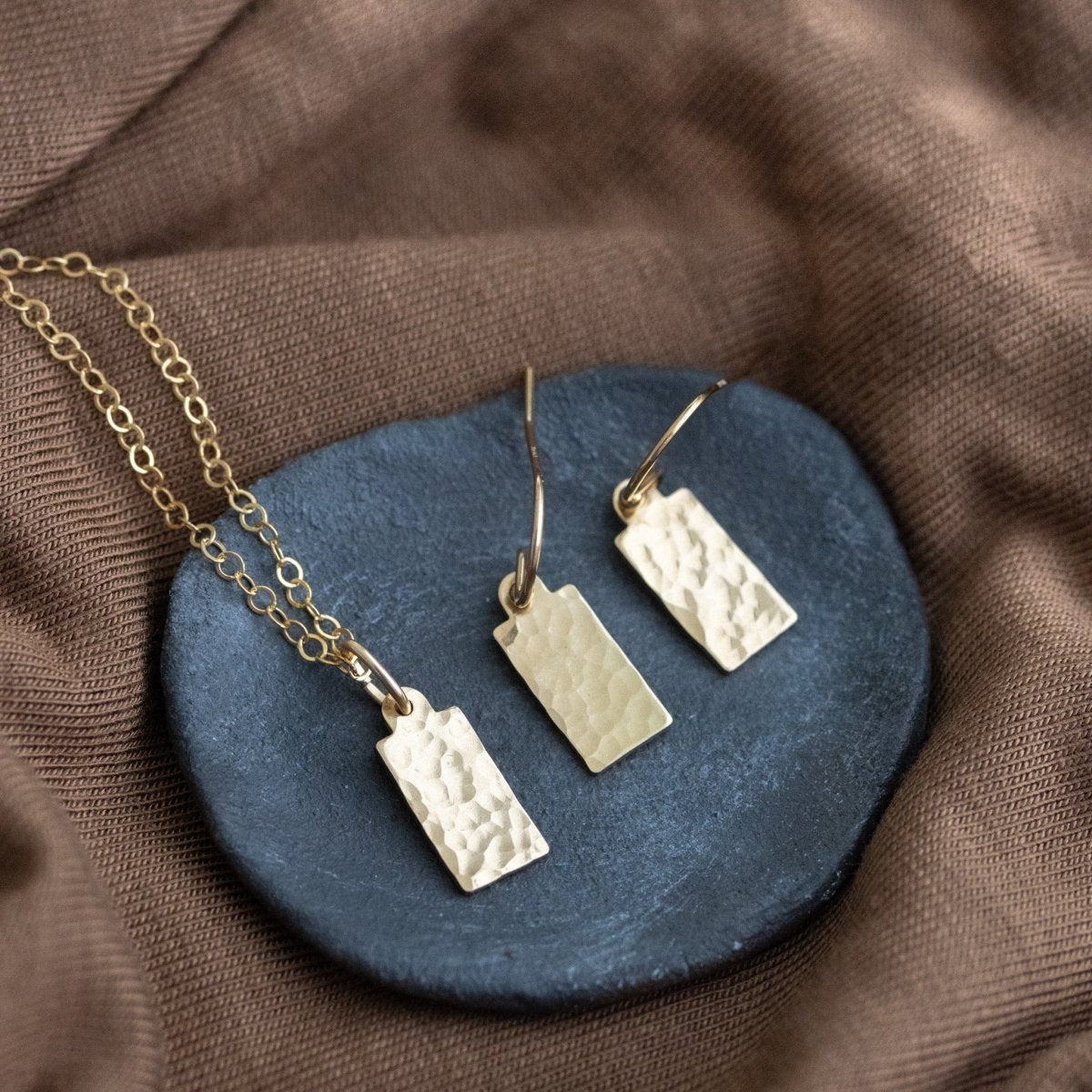 Eine Goldkette und ein Paar Ohrringe mit jeweils einem rechteckigen, gehämmerten Plättchen-Anhänger liegen auf einem grauen Steinschälchen, das auf braunem Stoff liegt.