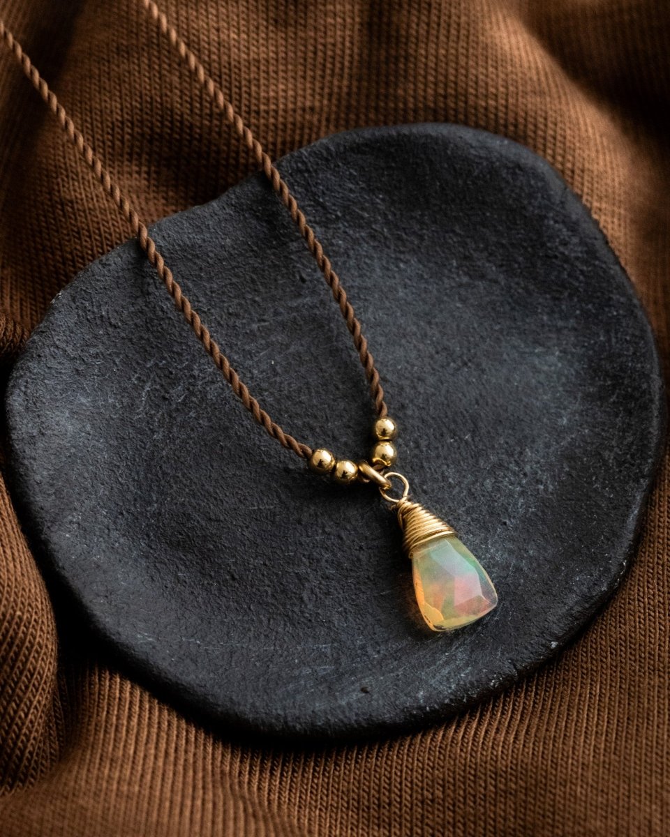 Eine dünne, braune Schnurkette mit einem dreieckigen Opal-Anhänger und goldenen Akzenten liegt auf einem schwarzen Schälchen.
