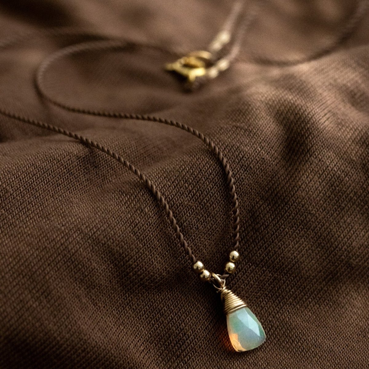 Eine dünne, braune Schnurkette mit einem dreieckigen Opal-Anhänger und goldenen Akzenten lehnt an braunem Stoff.