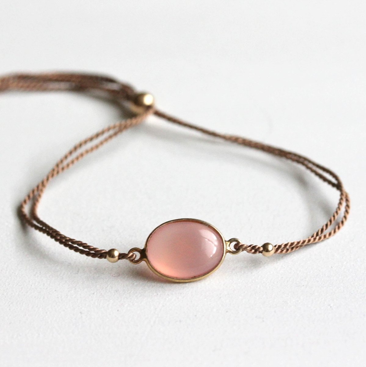 Ein beiges Seidenarmband mit einem ovalen, rosa Chalcedon im Zentrum auf weißem Hintergrund.