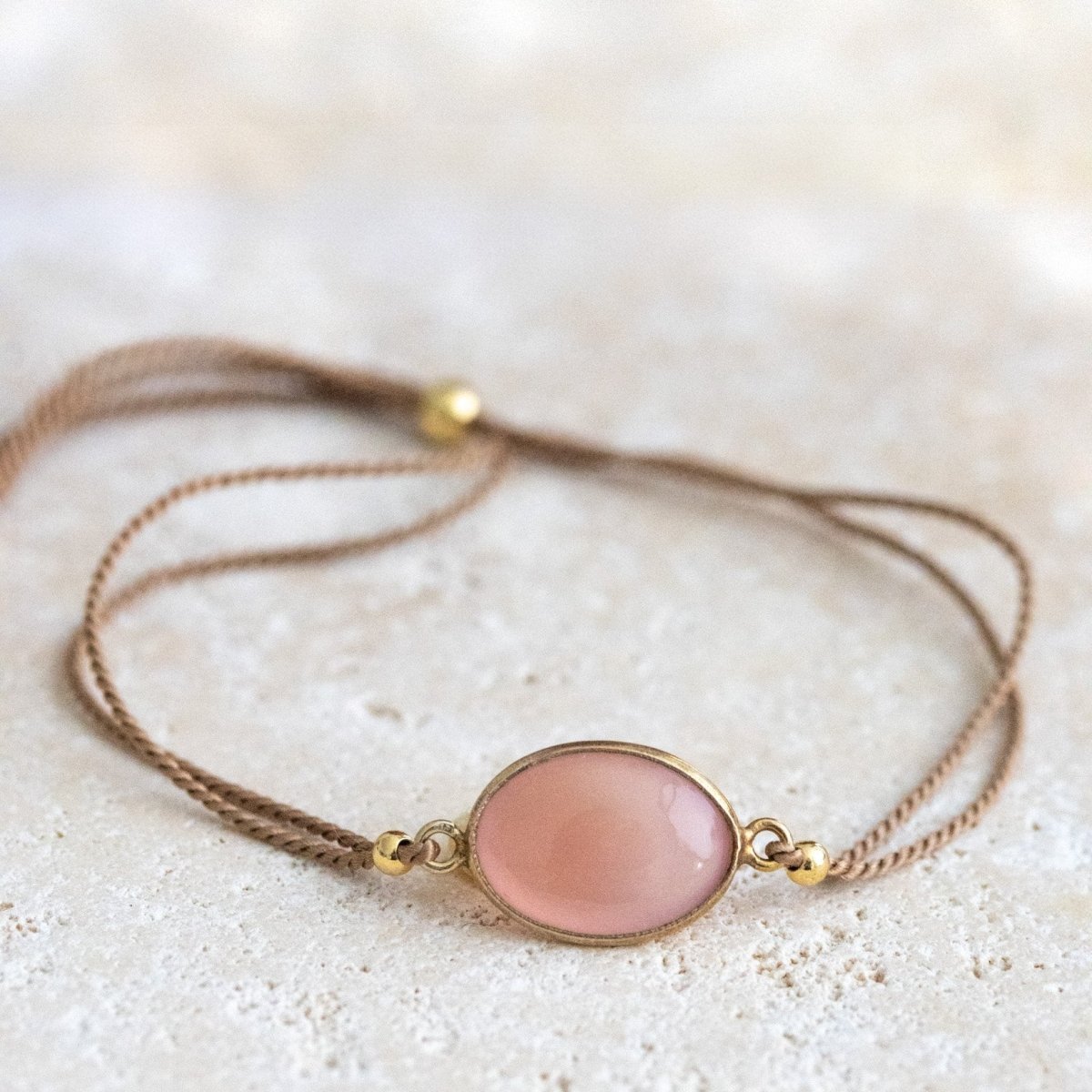 Ein beiges Seidenarmband mit einem ovalen, rosa Chalcedon im Zentrum auf einer hellen Natursteinplatte.