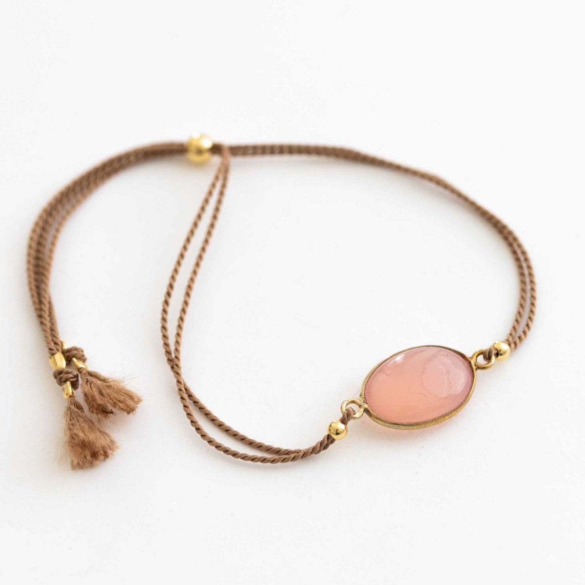 Ein beiges Seidenarmband mit einem ovalen, rosa Chalcedon im Zentrum auf weißem Hintergrund.