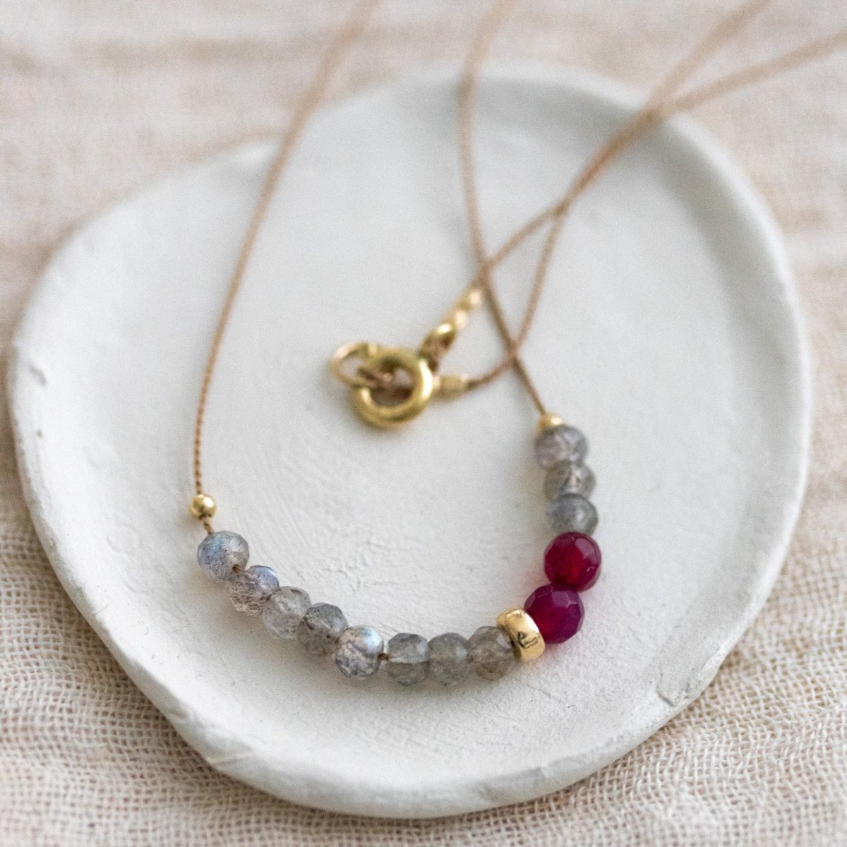 Eine Halskette aus beigem Seidenband, auf das einige Labradorit- und rote Achat-Rondelle aufgezogen sind, auf einem weißen Schälchen.