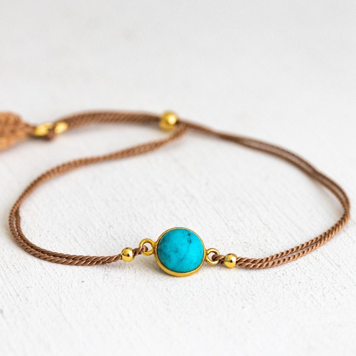 Ein dünnes, beiges Seidenkordel-Armband mit einem runden, gold gefassten Türkis-Edelstein in der Mitte, liegt auf weißem Hintergrund.