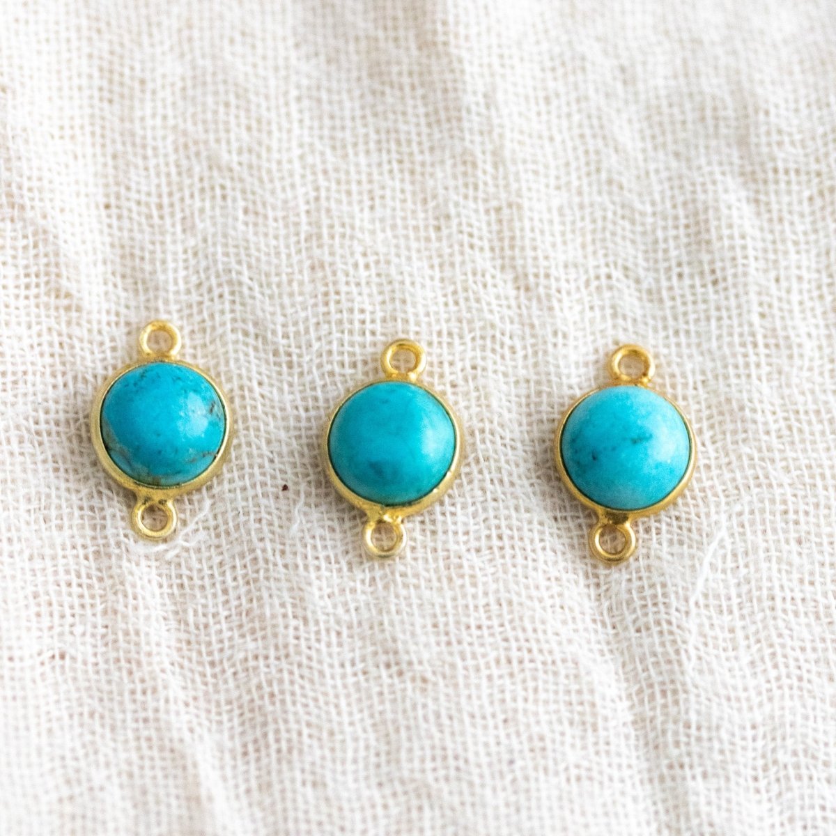 Drei runde und in gold gefasste Türkise liegen auf naturfarbenen Leinenstoff.