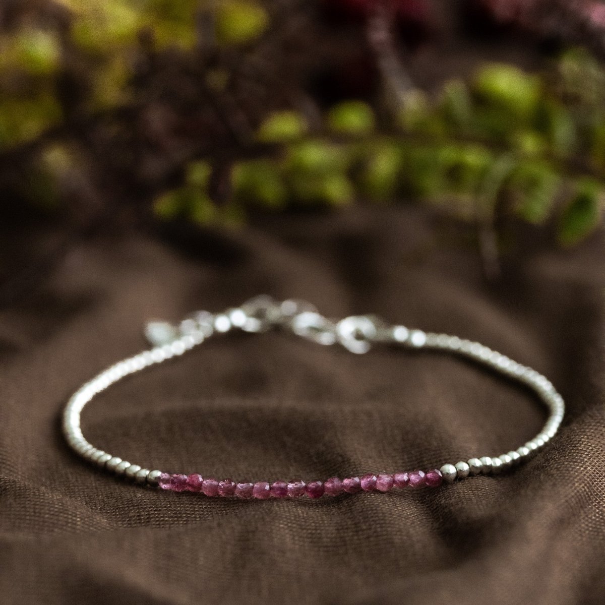 Ein sehr dünnes Silberarmband mit winzigen rosa Turmalinen in der Mitte liegt auf braunem Stoff. Verschwommener floraler Hintergrund.