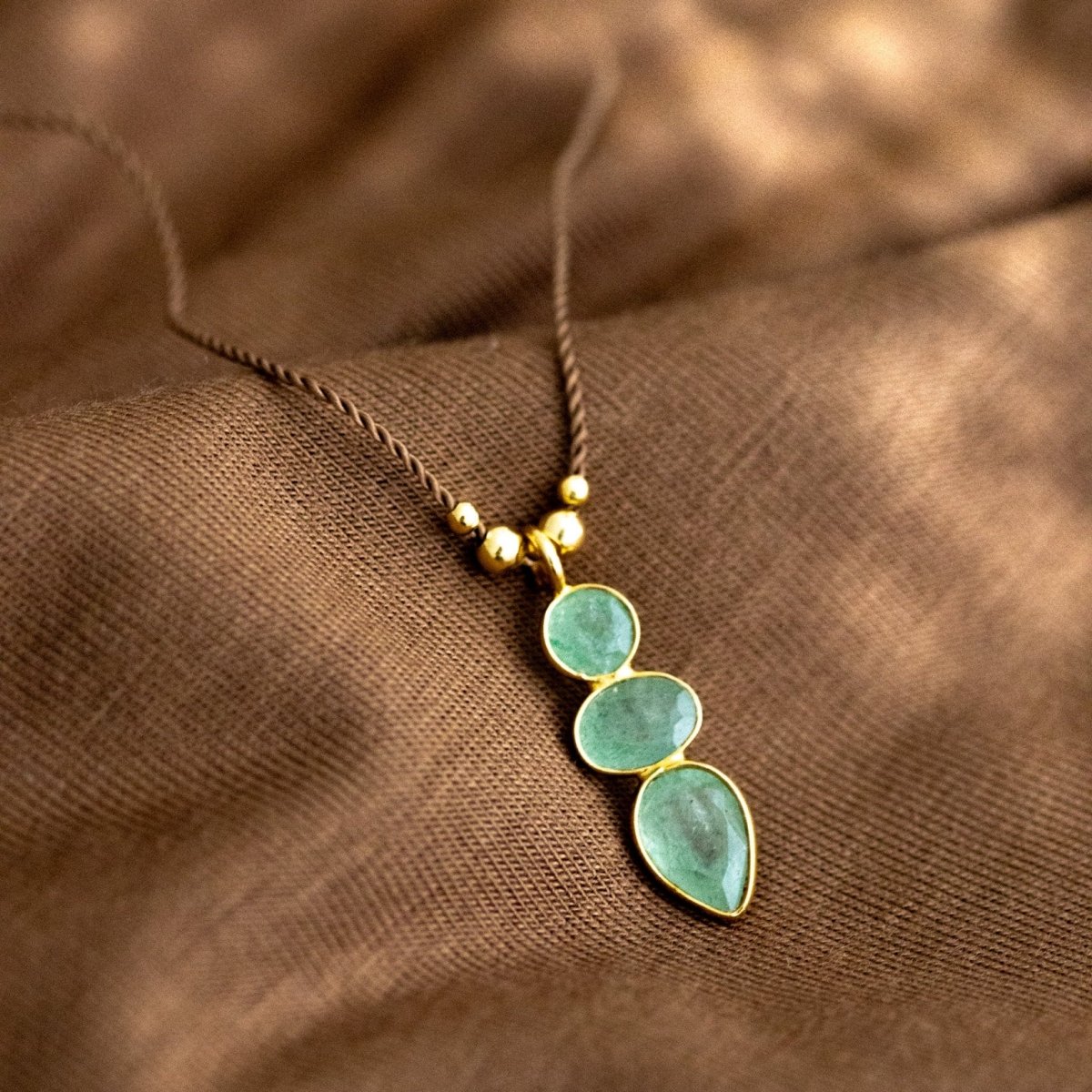 Eine lange, dünne Schnurkette mit einem länglichen Anhänger, der aus drei übereinander in gold gefassten Prehnit Edelsteinen besteht, liegt auf braunem Stoff.