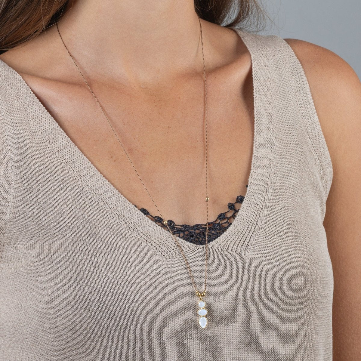 Eine lange Schnurkette mit einem länglichen Anhänger, der aus drei übereinander in gold gefassten Mondsteinen besteht, hängt am Hals des Models.