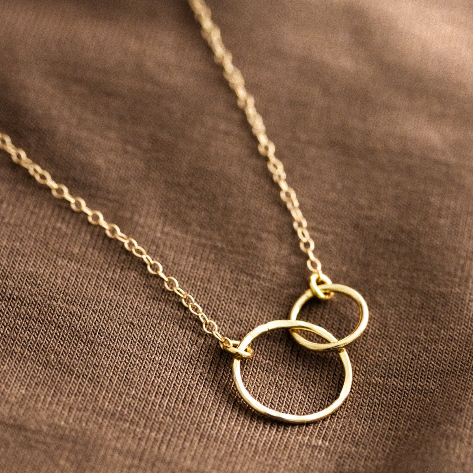 Eine feine Goldkette mit zwei ineinander verschlungenen Ringen in der Mitte liegt auf einem braunen Hintergrundstoff.