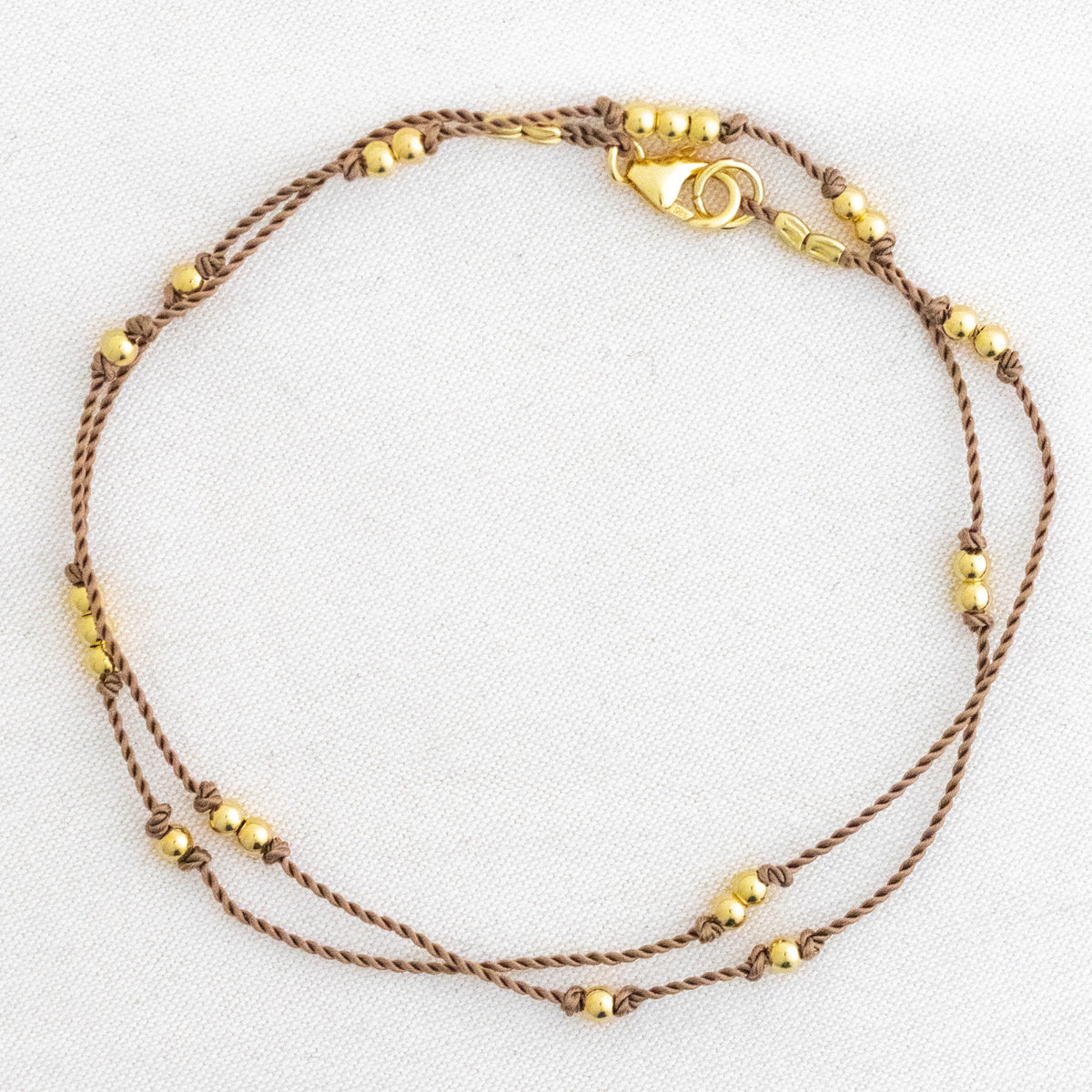 Dünne, beige Kordelkette mit unregelmäßig verteilten Goldperlen ausgelegt auf weißem Untergrund.