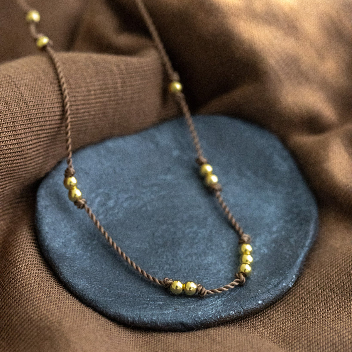 Eine dünne, beige Kordelkette mit unregelmäßig verteilten Goldperlen ausgelegt auf Schälchen.