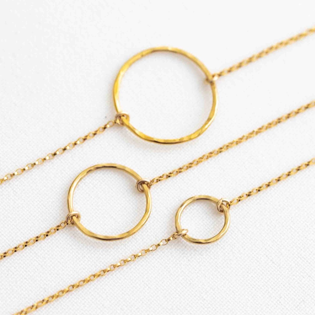 Drei feine, goldene Armbänder liegen auf weißem Hintergrund. Jedes der Armbänder hat einen offenen, gehämmerten Ring in der Mitte, bei jedem Armband in einer anderen Größe.