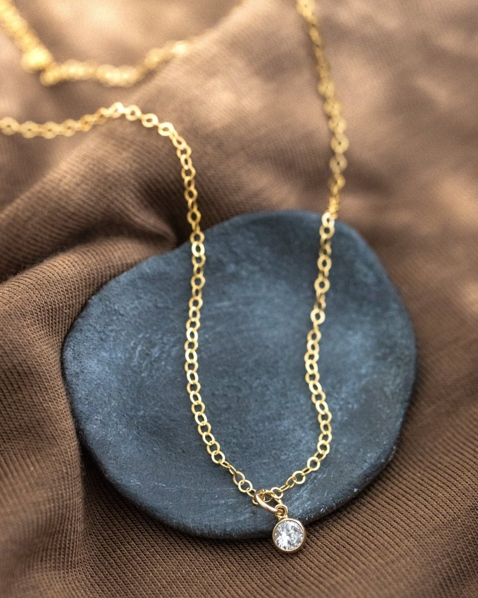 Eine feine Goldkette mit einem winzigen, runden Zirkonia-Anhänger lehnt an einem schwarzen Schälchen auf braunem Stoff.