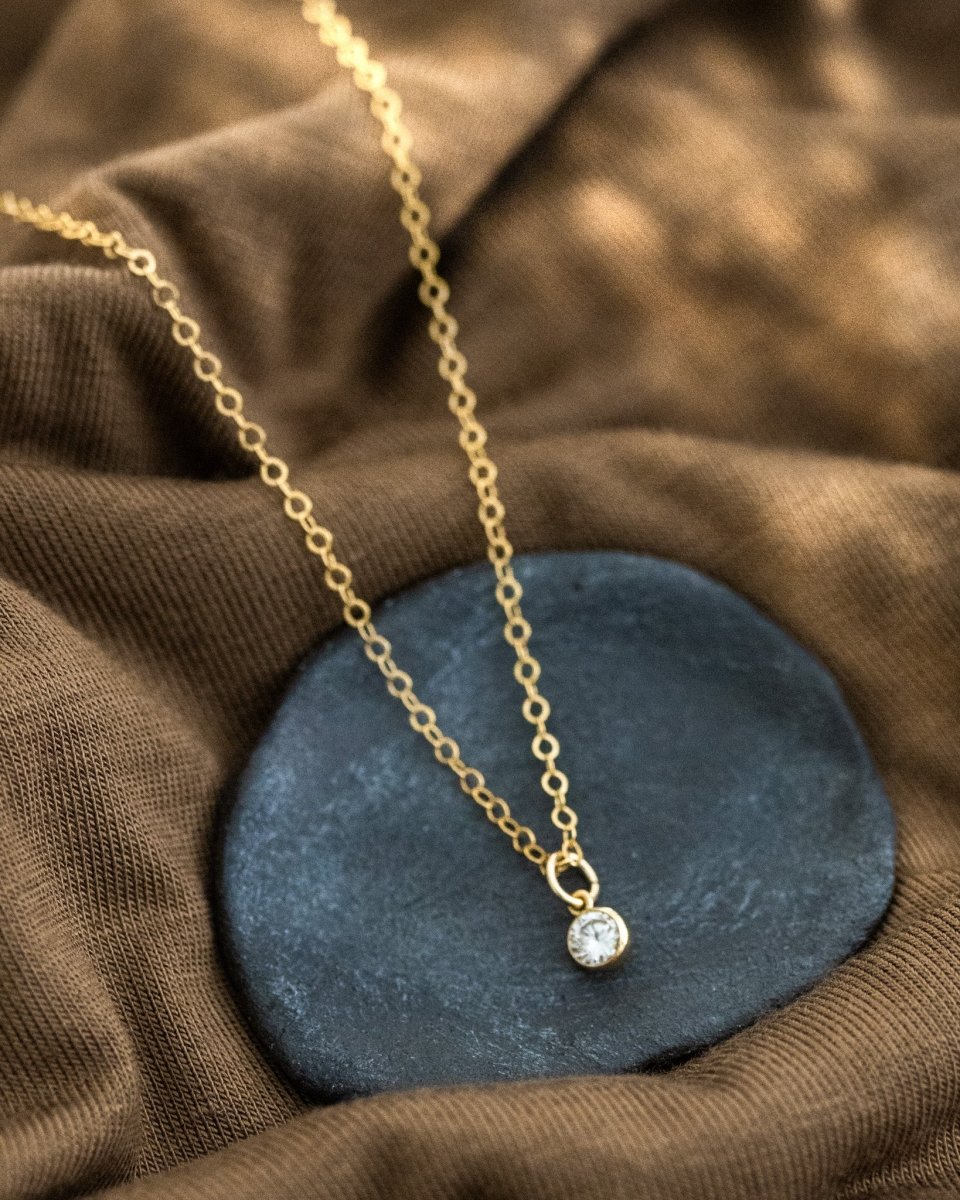 Eine feine Goldkette mit einem winzigen, runden Zirkonia-Anhänger liegt auf einem schwarzen Schälchen auf braunem Stoff.