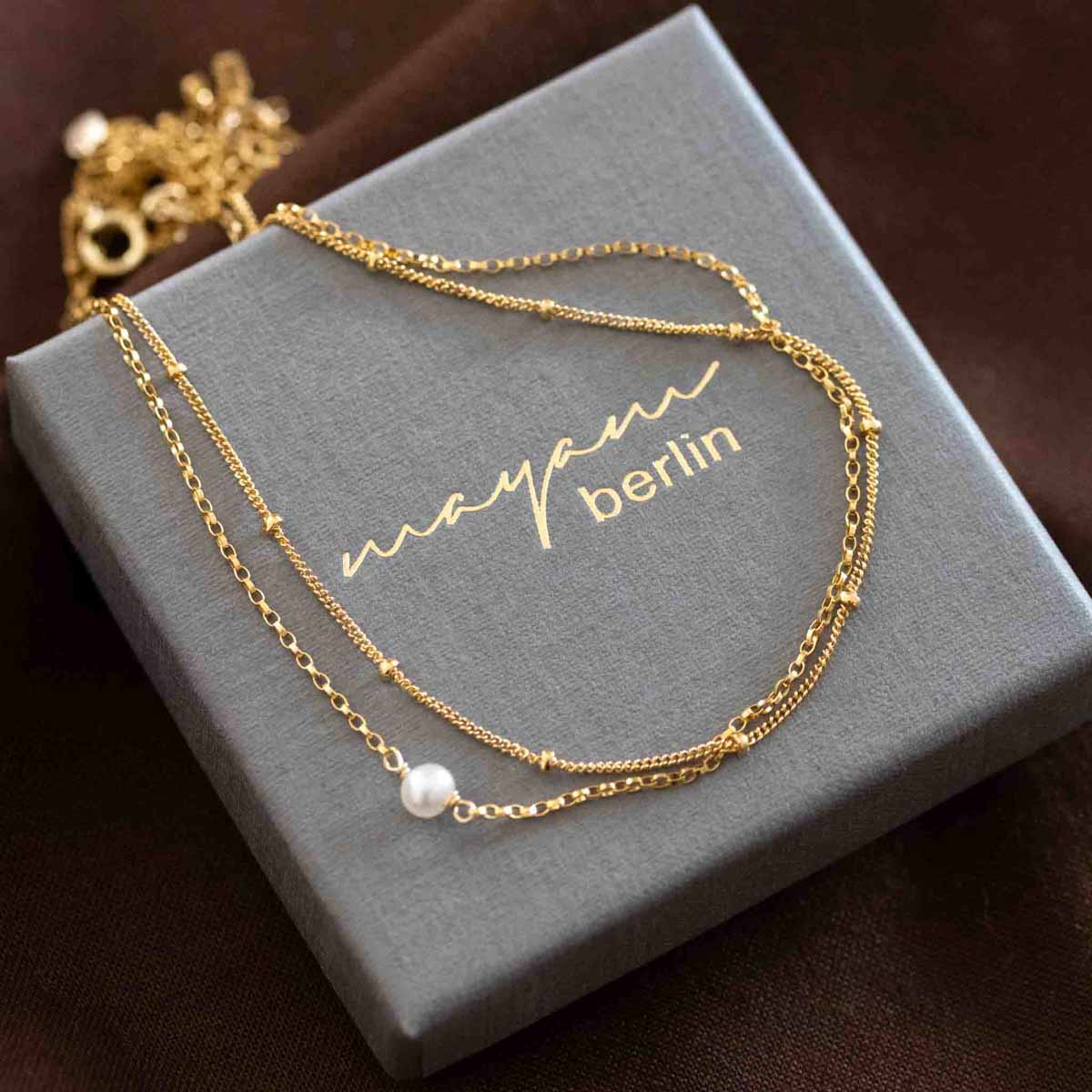Eine zweireihige Goldkette mit einer kleinen Süßwasserperle auf einem grauen Schmucketui.