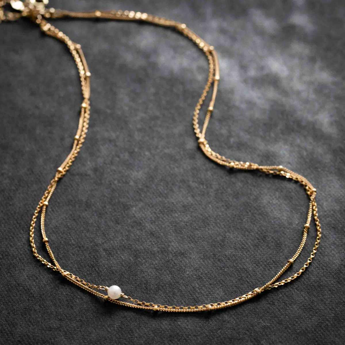 Eine zweireihige Goldkette mit einer kleinen Süßwasserperle auf schwarzem Hintergrund.