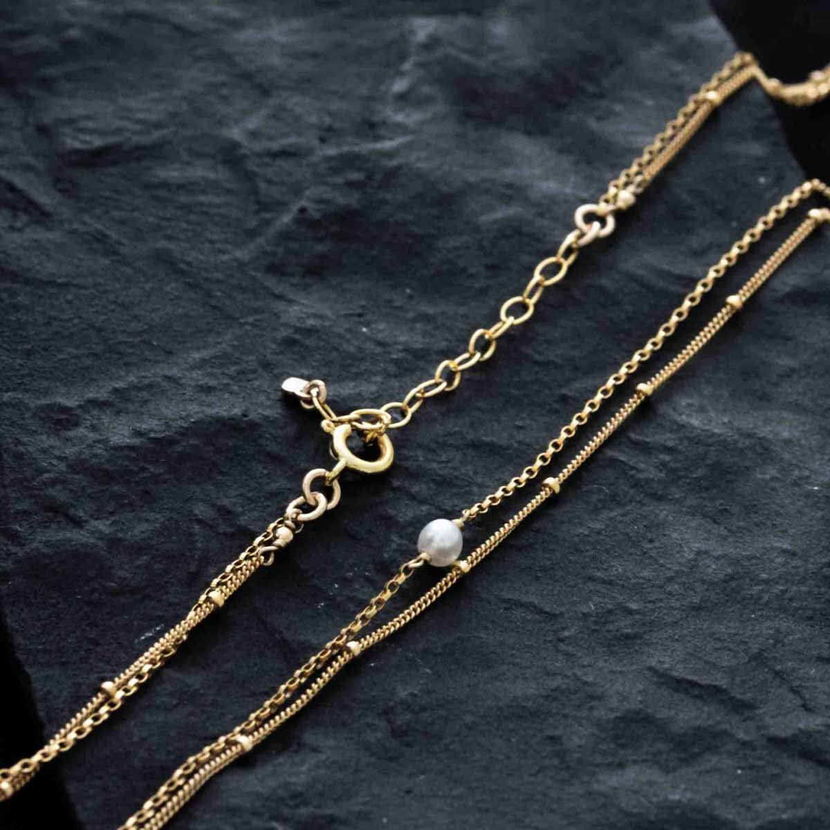 Eine zweireihige Goldkette mit einer kleinen Süßwasserperle auf schwarzem Hintergrund.
