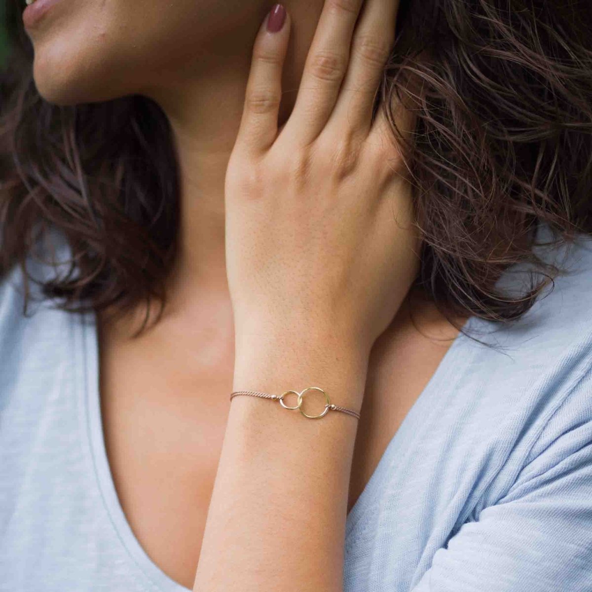 Am Handgelenk des Models hängt ein feines Seidenarmband mit zwei goldenen, ineinander verschlungenen Ringen in der Mitte.
