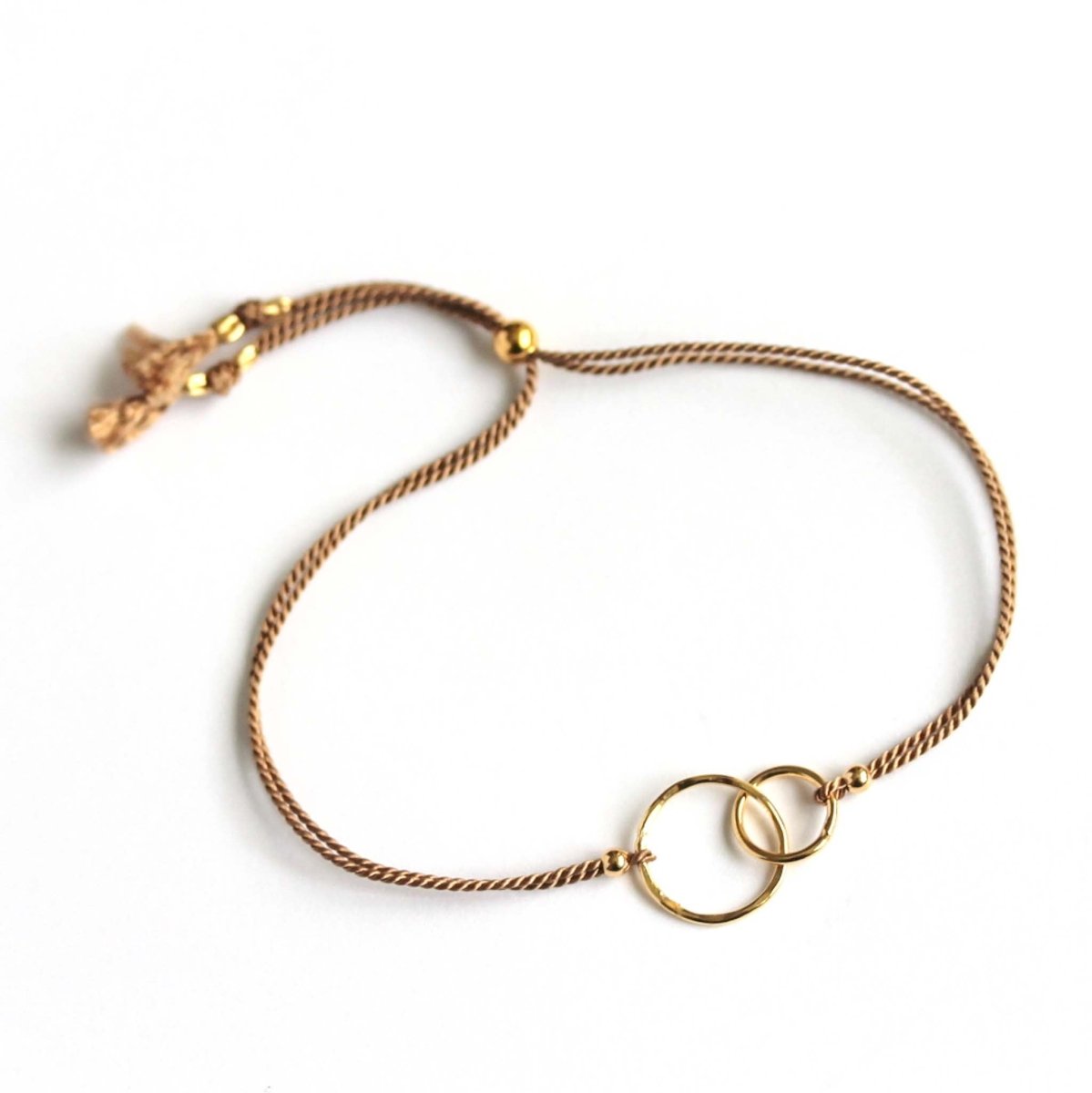 Ein beiges Seidenarmband mit goldenen, ineinander verschlungenen Metallringen in der Mitte, abgebildet auf weißem Hintergrund.